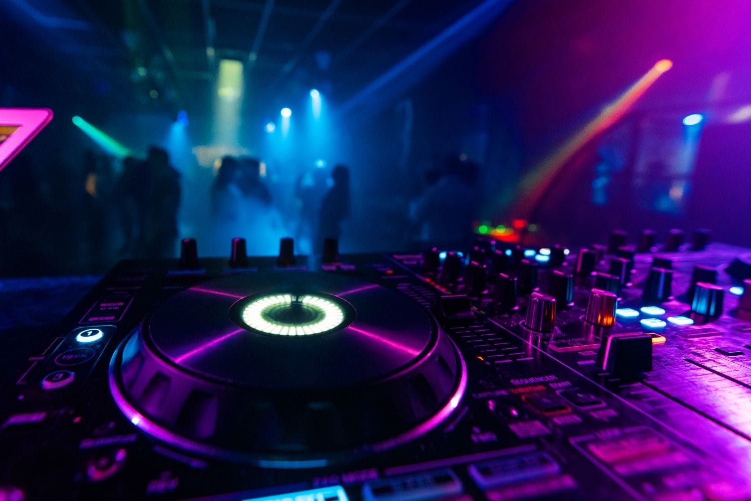 A DJ mixer glowing in the foreground with a hazy dancefloor and laser light behind it