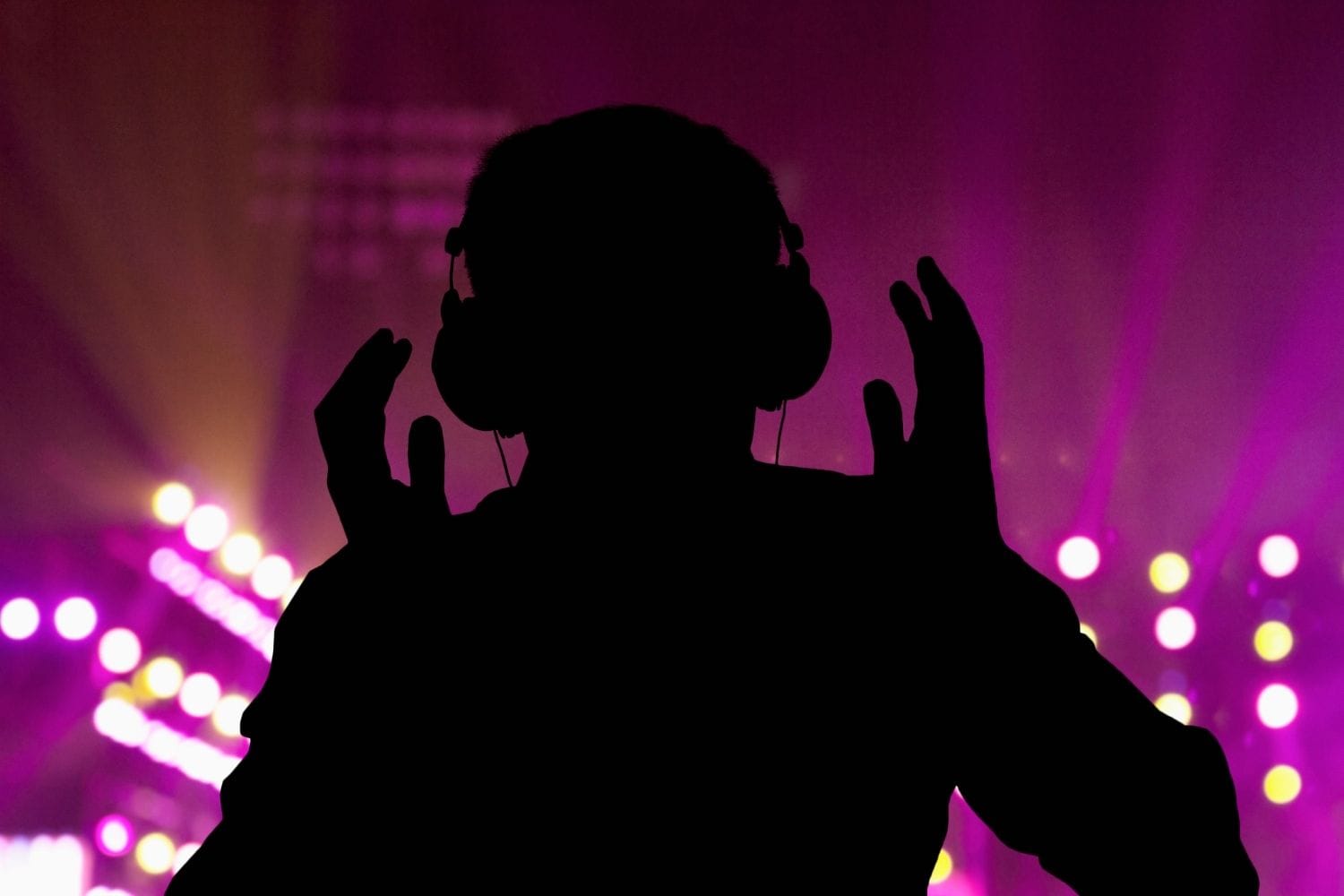 Silhouette of a listener with headphones raised in front of magenta stage lights and yellow bokeh