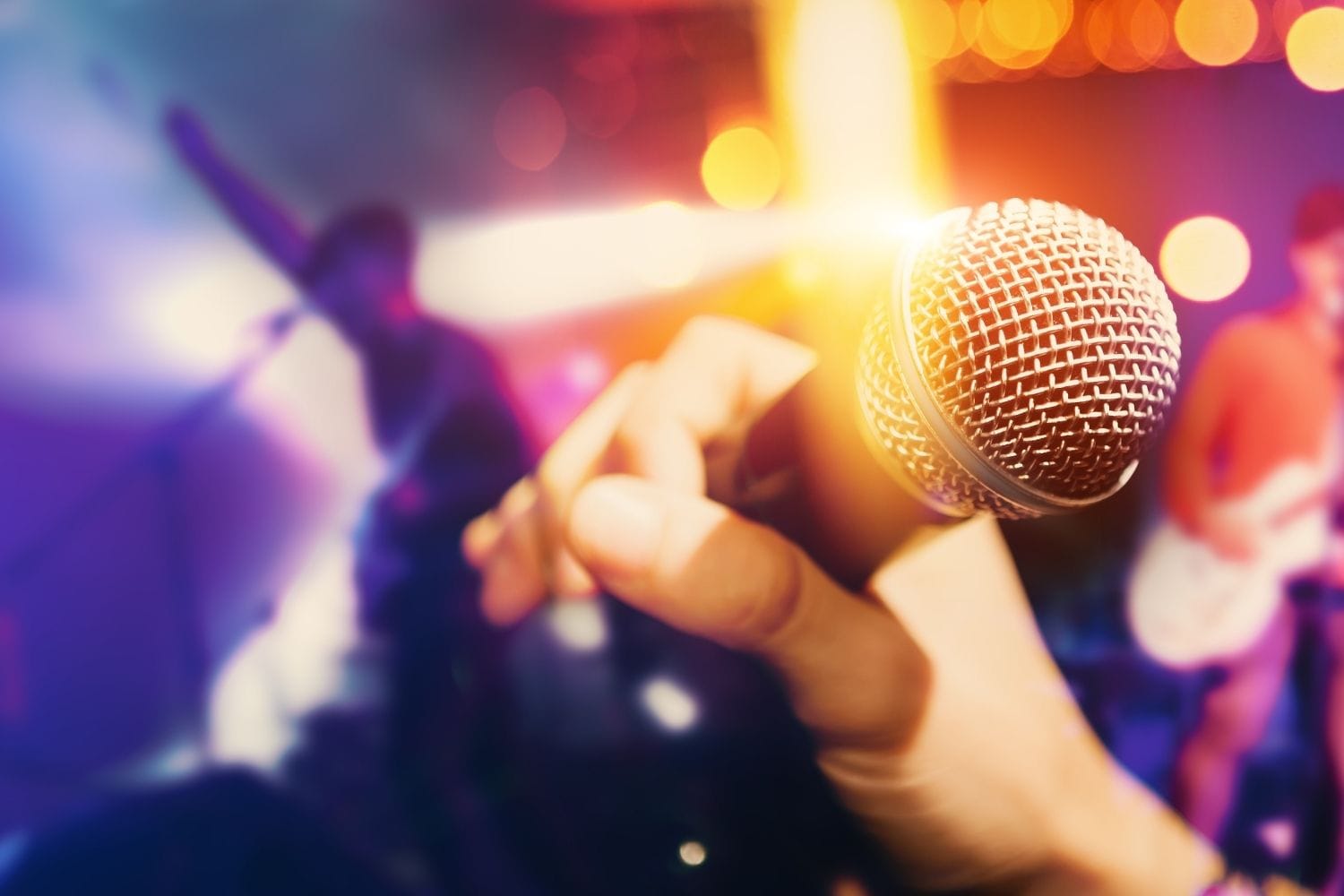 A hand holding a microphone up on a lit stage with warm bokeh behind