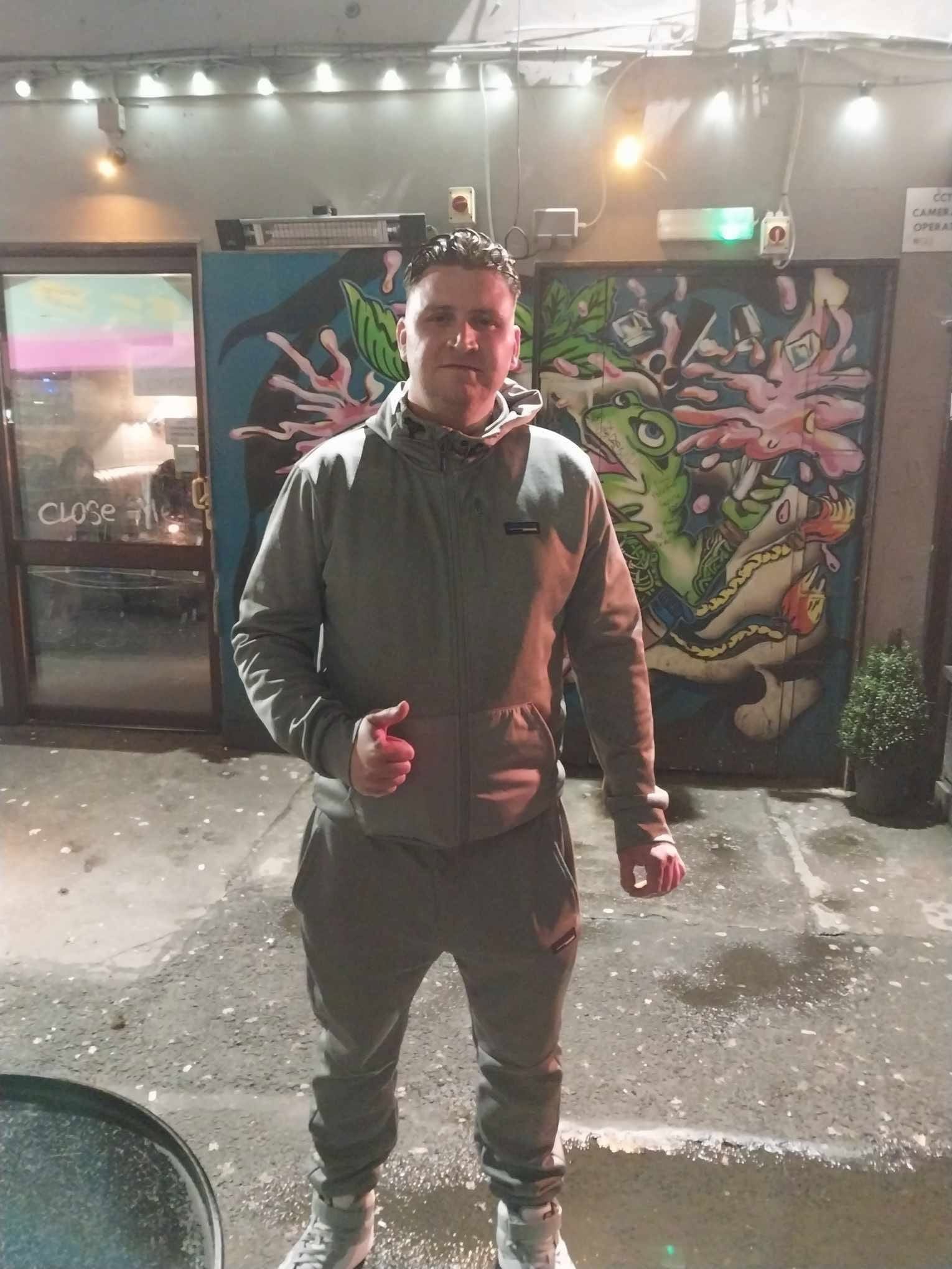 Brian M in a grey tracksuit outside a venue, in front of a painted mural