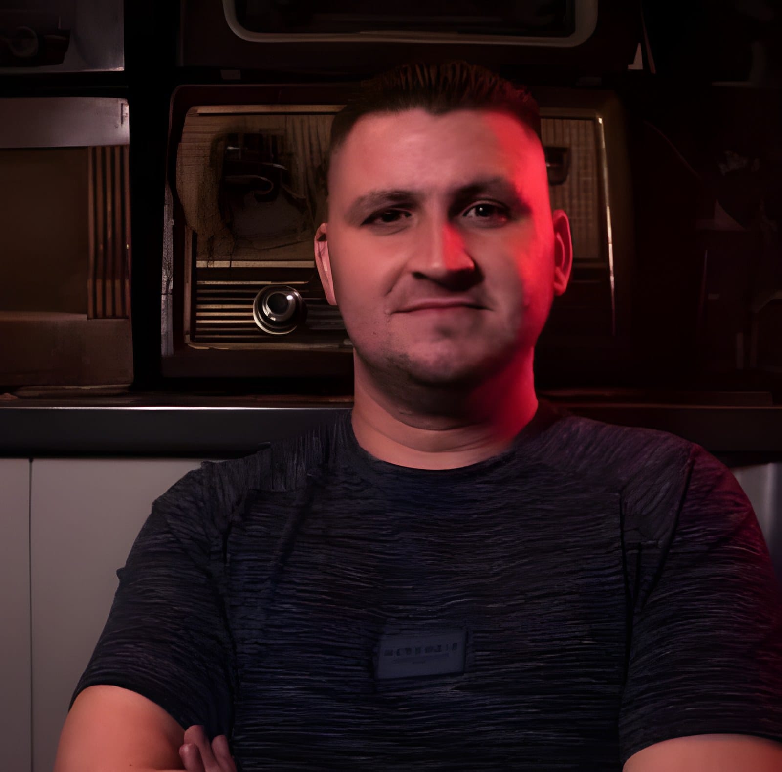 Brian M sitting with arms folded in front of a shelf of vintage radios, lit in red